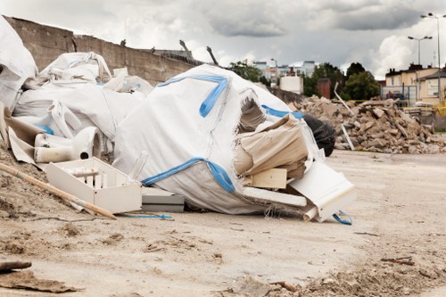 A large pile of construction and rubbish waste is situated on a dirt surface within a construction site or outdoor area, consisting of white plastic sheeting, rolled-up pipe sections, and various discarded materials. The waste appears loosely piled, with some items partially collapsed or leaning. In the background, there is a partially demolished wall or barrier, along with distant industrial and residential buildings, trees, and a cloudy sky overhead. To the right, there is a mix of rubble and broken concrete, indicating ongoing building or demolition work. The scene is indicative of a typical rubbish removal scenario managed by a waste clearance service such as Garden Clearance Wallington, which operates in the local area around Wallington, London. The image underscores the importance of professional waste disposal and clearance services for construction and site tidy-up projects in the region.