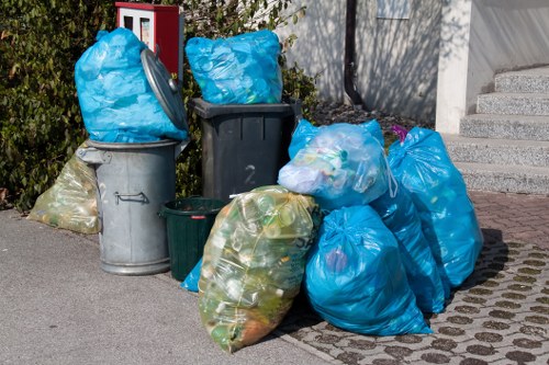 A collection of several large refuse bags, predominantly blue, filled with various garden and household waste, are placed on a paved driveway outside a residential property. The bags are situated next to a black and a grey wheelie bin, with additional smaller green and transparent bags containing mixed waste stacked nearby. The black bin appears empty or partially filled, while the grey bin and blue bags are overflowing with waste materials. The surrounding environment includes a concrete staircase and a wall of a residential building, with some greenery visible to the left, suggesting a garden clearance area. The scene reflects typical rubbish disposal for garden or household waste collection, consistent with services offered by Garden Clearance Wallington in the local TW postcode area. The overall setting is well-lit, indicating daytime, with no visible signs of recycling or selective waste sorting in this particular collection. This arrangement is typical of rubbish removal and garden clearance processes in the local area.