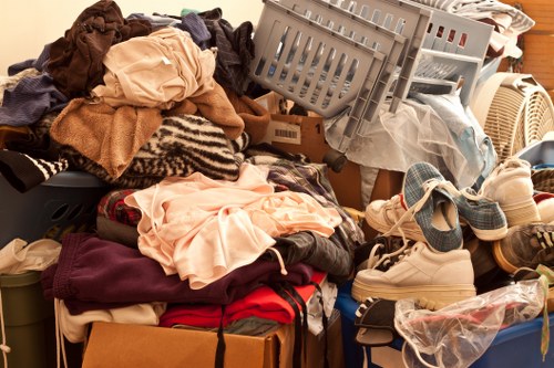 A large pile of mixed household waste and clutter, including clothing, textiles, and personal items, is seen on a wooden floor or surface. The clothing consists of various fabrics such as cotton, wool, and synthetic materials, in multiple colours including beige, brown, red, and black, some folded and others loosely piled. Among the textiles are folded shirts, sweaters, and scarves, with some items partially crumpled or draped over the pile. Visible footwear, including a pair of sports shoes and slippers, rests on or near the clutter. There are also plastic bags, cardboard boxes, and a gray plastic laundry basket tilted on its side, containing more discarded items. The environment appears to be indoors, possibly a storage room or corner of a home, with surrounding objects including a radiator and a wooden panel wall. This scene illustrates waste accumulation that a professional rubbish removal service like Garden Clearance Wallington might handle for homeowners needing garden clearance or household decluttering within the Wallington area.