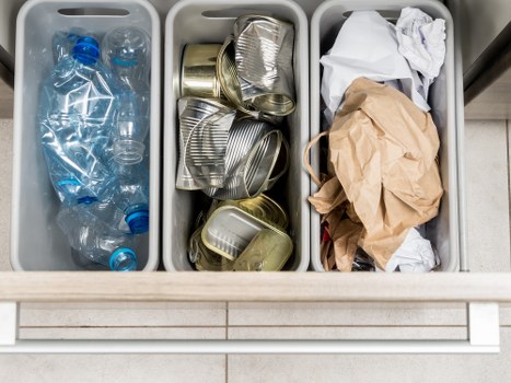 An image showing three grey plastic bins on a tiled floor, each containing different types of waste suitable for rubbish removal services by Garden Clearance Wallington. The left bin holds several clear plastic water bottles, some with open caps and labels, indicating recyclable drink containers. The middle bin contains stacked and slightly crushed metallic cans, primarily aluminium, with some showing ridged surfaces typical of beverage cans. The right bin is filled with crumpled and loose paper waste, including a brown paper bag and white paper scraps, suggesting waste material ready for collection or disposal. The bins are positioned side by side on a paved surface, with the scene illuminated by natural light, emphasizing the textures and colors of the waste materials, which range from transparent plastics to shiny metallic cans and Kraft paper. This setup reflects a typical rubbish collection scenario for domestic or small business waste transfer, aligned with services provided by Garden Clearance Wallington in close proximity to the town's local waste management infrastructure.