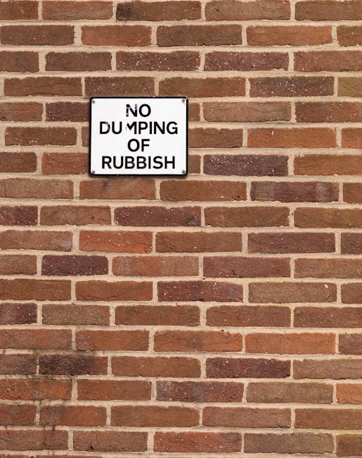 A rectangular metal sign mounted on a red and brown brick wall displays a message in black capital letters that reads 'NO DUMPING OF RUBBISH,' with the words arranged in four lines. The sign has a whi