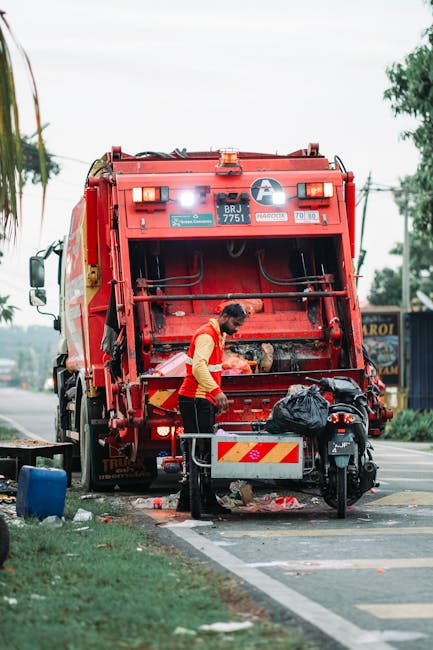 A small, light blue pickup truck with a black cargo bed is parked alongside a street, overloaded with various types of rubbish and debris. The truck's body shows signs of wear and rust, with visible s